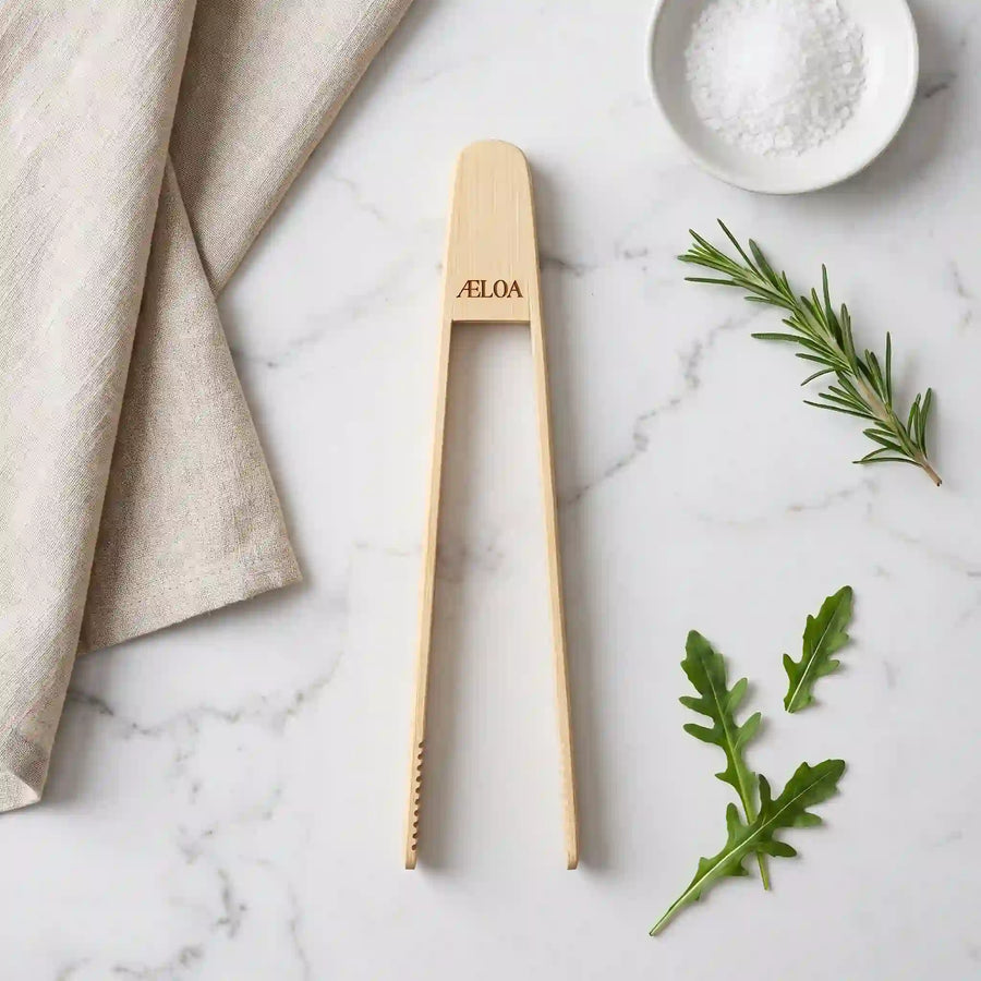 Madpincet i bambus med indgraveret ÆLOA-logo set oppefra på marmor med hørklæde, havsalt, rosmarin og rucola