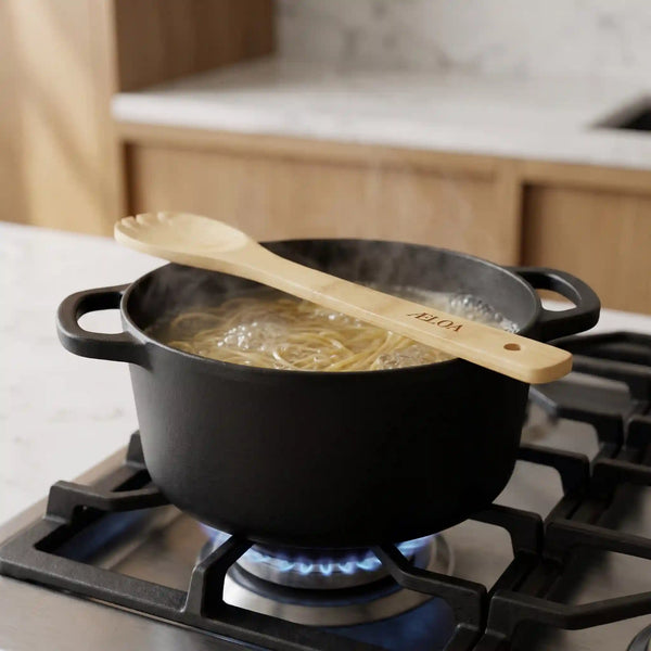 Pastaske i bambus med indgraveret ÆLOA-logo hviler på støbejernsgryde med kogende spaghetti på gaskomfur